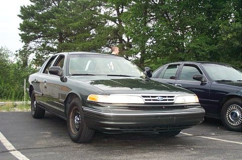 1991 Ford LTD Crown Victoria - A dark green Ford Crown Victoria is parked in a lot next to another vehicle, with trees in the background.
