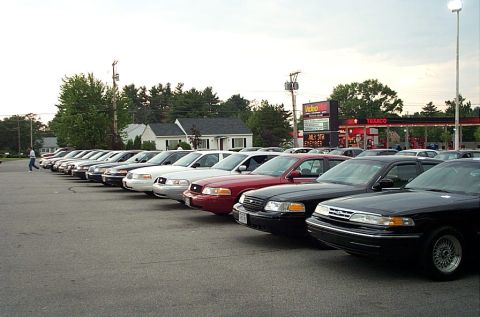 1991 Ford LTD Crown Victoria - A row of Ford Crown Victoria sedans are parked in a lot, with a gas station visible in the background.