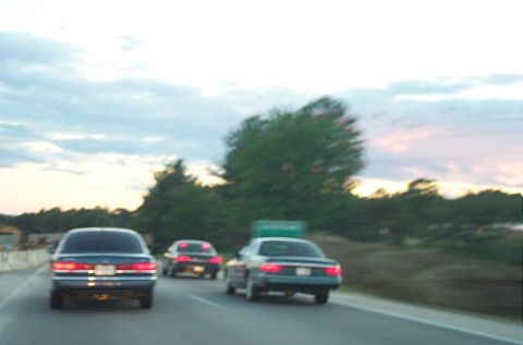 1991 Ford LTD Crown Victoria - Three cars drive down a highway under a cloudy sky at dusk.