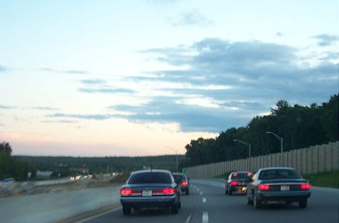 1991 Ford LTD Crown Victoria - Several cars travel down a highway as the sun sets, casting a soft glow on the clouds.