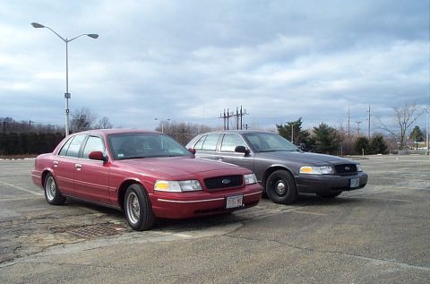 1991 Ford LTD Crown Victoria - Two Ford Crown Victoria sedans, one red and one gray, are parked in an empty lot under a cloudy sky.