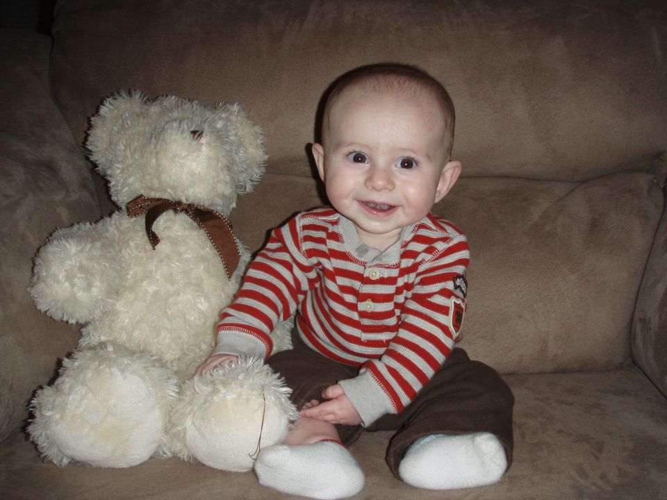Baby in striped shirt and brown pants sits next to a large white teddy bear on a brown couch