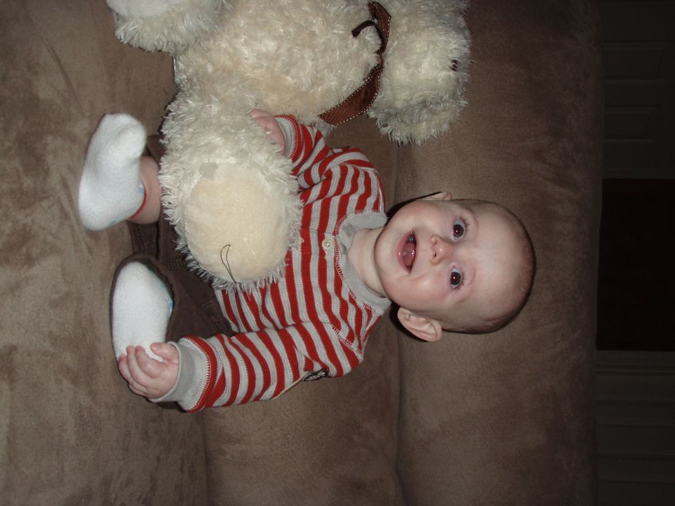Baby in a striped shirt and white socks holding a teddy bear on a brown couch