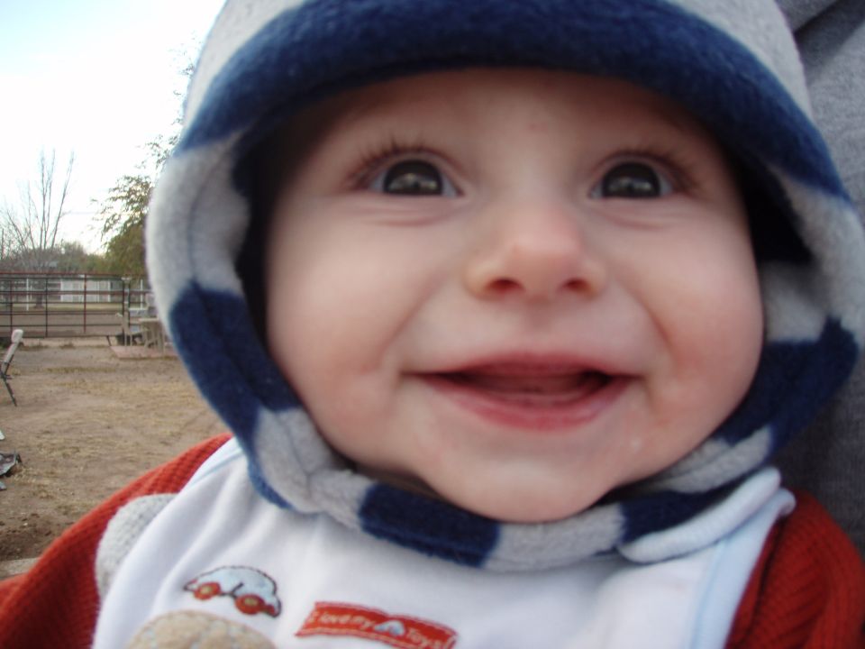 Baby in a blue and gray striped hood, smiling. There's a fence and some trees in the background