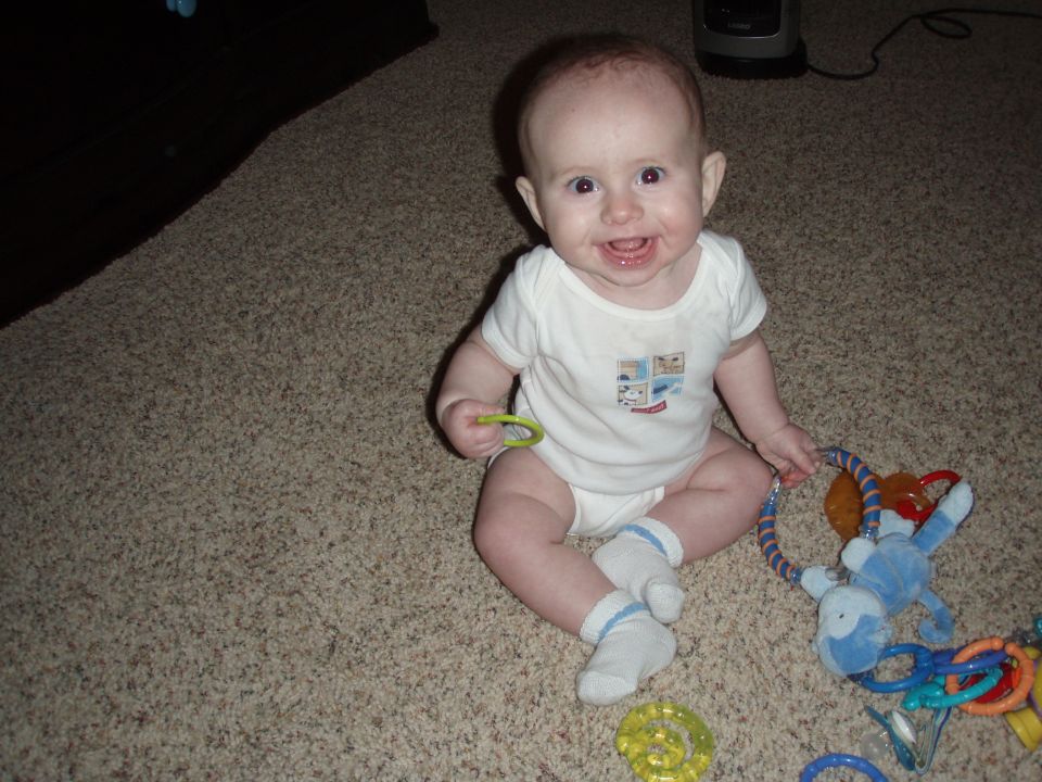 Baby sitting on carpet with toys, Supermotors logo visible