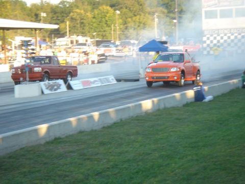 2007 Ford Mustang - Two pickup trucks compete on a drag strip, with smoke trailing from the orange truck.