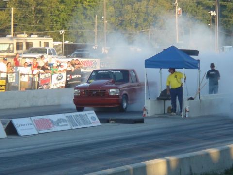 2007 Ford Mustang - A red pickup truck performs a burnout at a drag racing event, creating a large cloud of smoke.