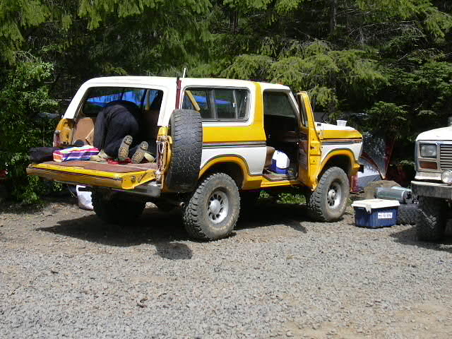 Yellow and white Ford Bronco with rear tailgate down and spare tire, gear scattered at a gravel campsite