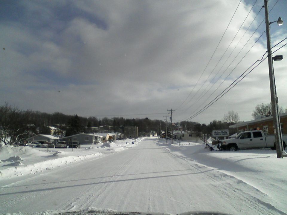 Snowy street scene with houses, power lines, and a white pickup truck with a snowplow attached. The truck is parked in front of a building with a sign