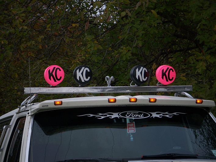 Top of a Ford truck with a light bar and four KC lights, two pink and two black. Flames decal on the windshield
