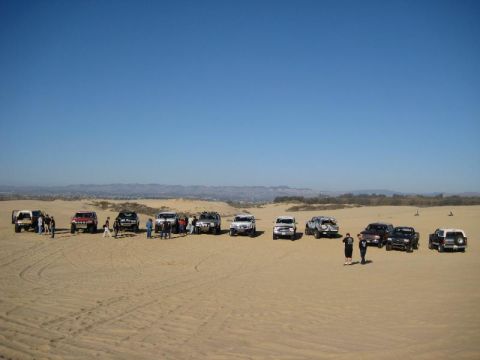 1989 Ford Bronco - A group of SUVs and trucks are parked on sand dunes with a distant city and mountains in the background.