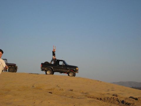 1989 Ford Bronco - Two people stand atop a black 1989 Ford Bronco on a sandy dune, with another vehicle nearby.