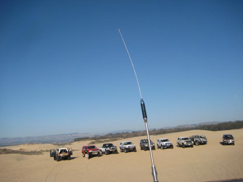 Several SUVs and trucks parked in sand dunes under a clear blue sky. An antenna is in the foreground