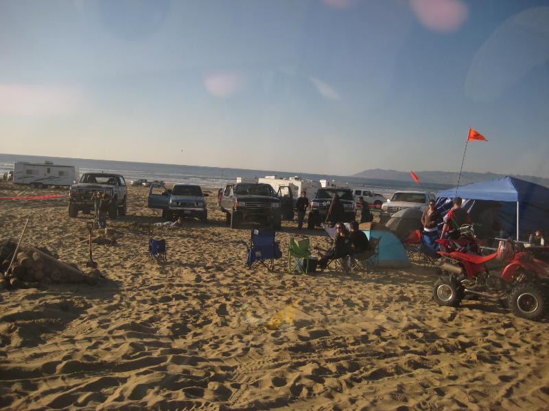 Beach scene with several trucks, RVs, tents, and an ATV on the sand under a clear sky
