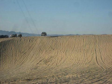 1989 Ford Bronco - Multiple vehicles traverse sandy terrain, leaving tracks on the dunes under a vast blue sky.