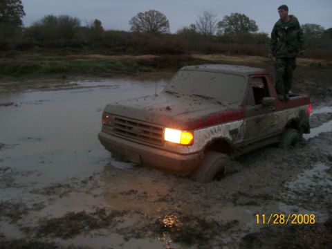 1988 Ford Bronco - A person stands atop a mud-covered pickup truck that is deeply stuck in a muddy, watery area.