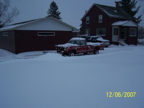 1989 Ford F-150 - A red pickup truck equipped with a snow plow sits in a snow-covered driveway, with a house and garage in the background.