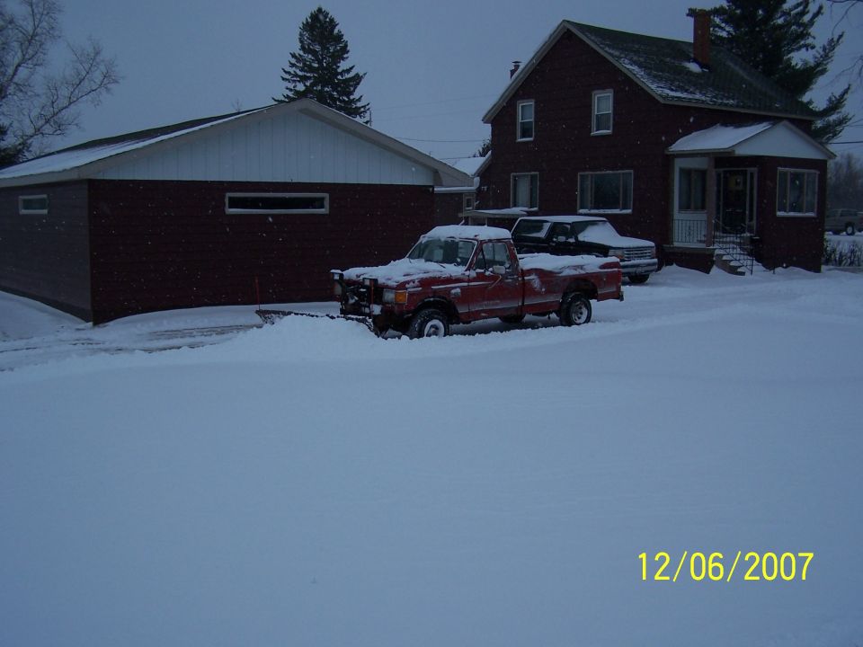 Red pickup truck with a snow plow attached, parked in a snowy driveway next to a garage and house. Another vehicle is parked behind it