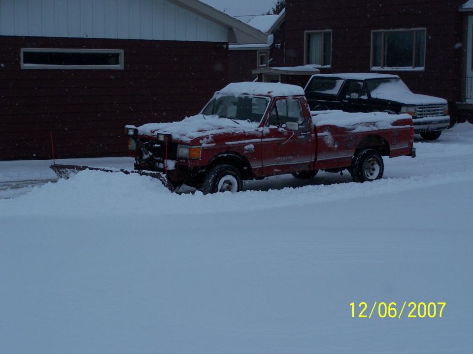 Red 1989 Ford F-150 pickup truck with a snow plow clearing snow, parked in front of a house
