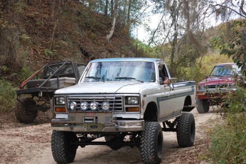 1982 Ford F-100 - Two lifted pickup trucks navigate a dirt path through a wooded area.