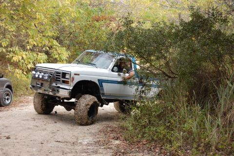 1982 Ford F-100 - A lifted white pickup truck with a blue stripe navigates a dirt path through trees, with another vehicle visible to the left.