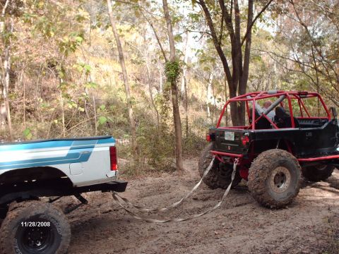 1982 Ford F-100 - A 1982 Ford F-100 pickup truck is seen from the rear, being towed by a custom off-road vehicle in a wooded area.