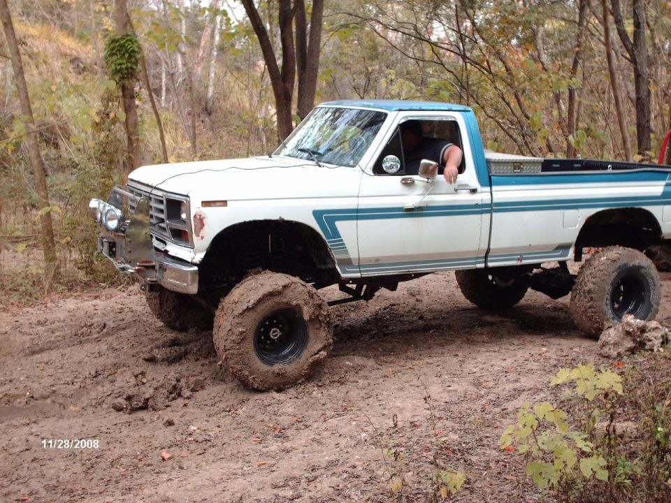 White and blue pickup truck with large tires driving through mud. Chrome bumper and side trim visible