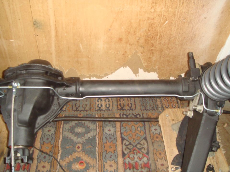 Black differential axle assembly with coil spring and brake lines against a patterned rug and textured wall