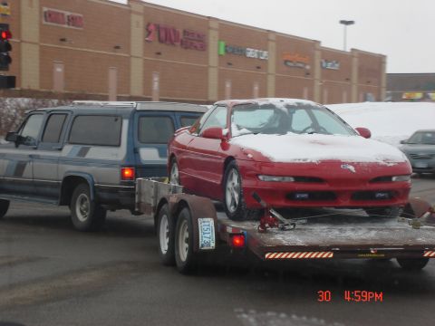 1993 Ford Probe - A red 1993 Ford Probe is transported on a trailer, partially covered in snow, with a blue SUV towing it.