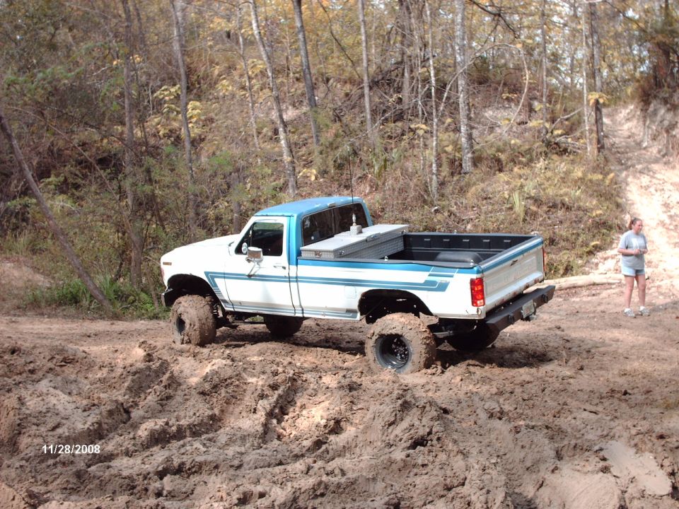 White and blue Ford pickup truck with lifted suspension driving through mud. Toolbox in bed