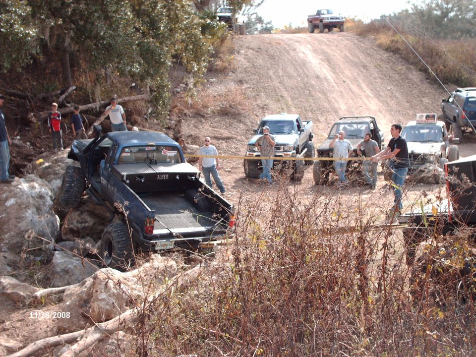 Several off-road trucks are navigating a rocky, dirt trail with people watching