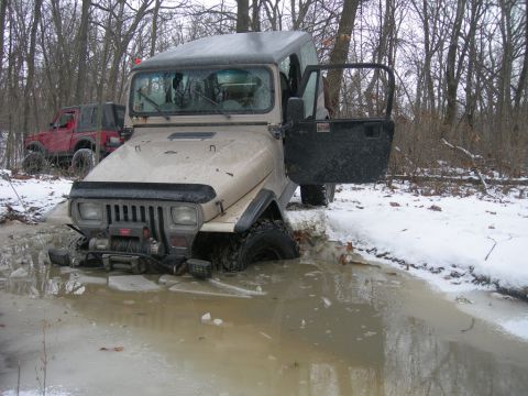 1991 Jeep Wrangler - A tan 1991 Jeep Wrangler is stuck in icy water in a wooded area, with its driver's door open. Another red Jeep is visible in the background.