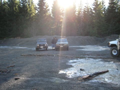 1980 Ford Bronco - Multiple SUVs are parked in an outdoor area with trees and gravel under a bright, glaring sun.