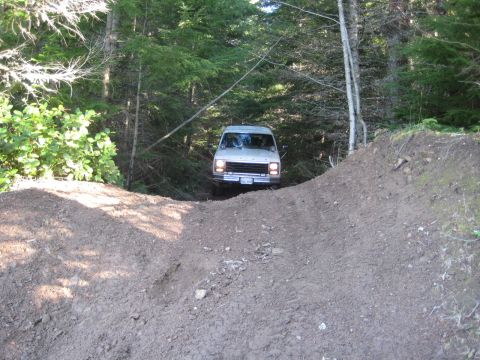 1980 Ford Bronco - Van navigates a rough dirt track through a forest, with a steep embankment in the foreground.
