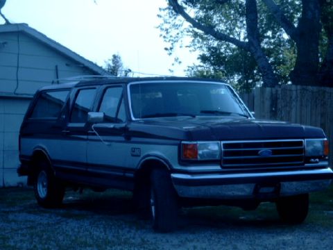1990 Ford Bronco - This Ford Bronco SUV is parked in front of a building with trees and a fence in the background.