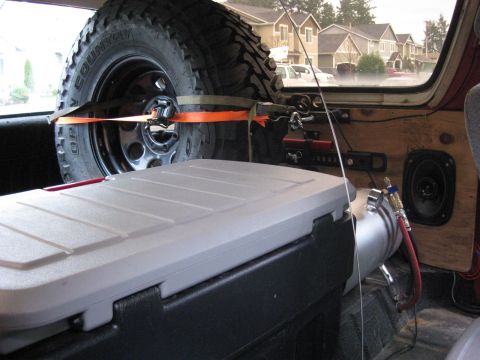 1979 Ford Bronco - Interior view of a vehicle showing a spare tire and cooler, with a suburban street scene outside.