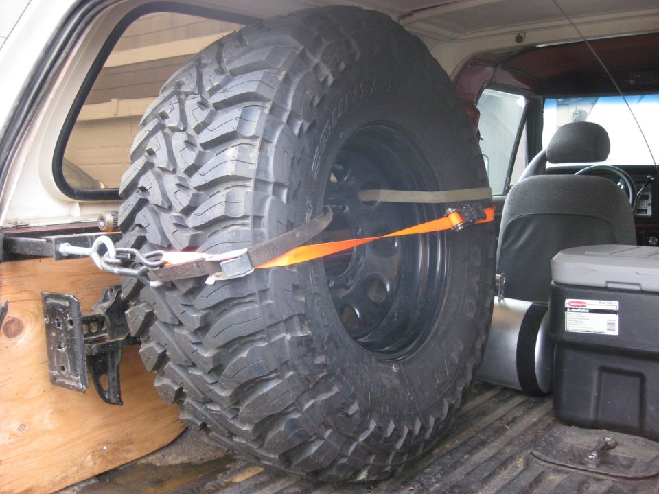 Spare tire secured with straps inside a vehicle's cargo area, showing part of the interior and a storage box