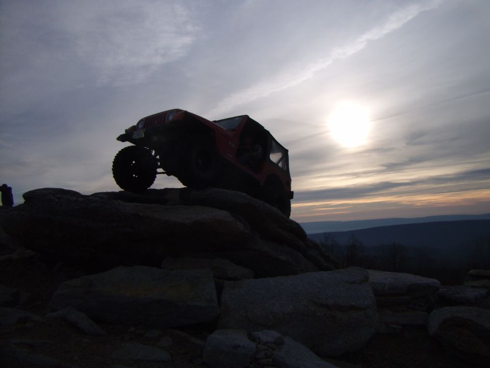 Red 2003 Jeep Wrangler climbing large rocks with sun setting in the background