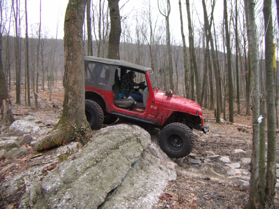 Red Jeep Wrangler navigating a rocky, wooded trail. The vehicle is angled towards the viewer's left, with its front right tire on a large rock