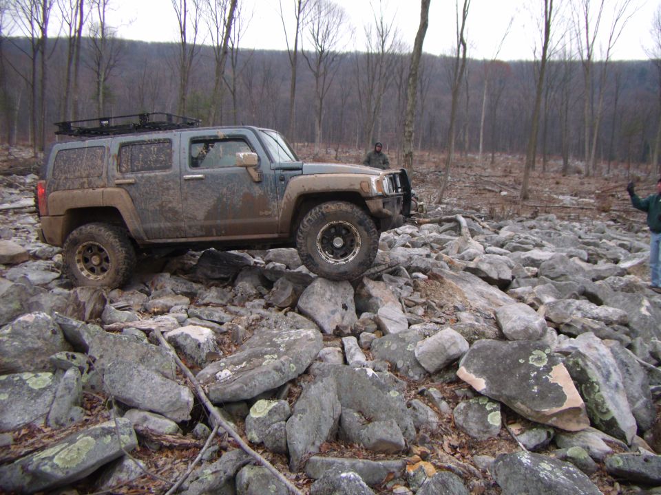 Mud-covered Hummer H3 navigating a rocky trail, trees in background