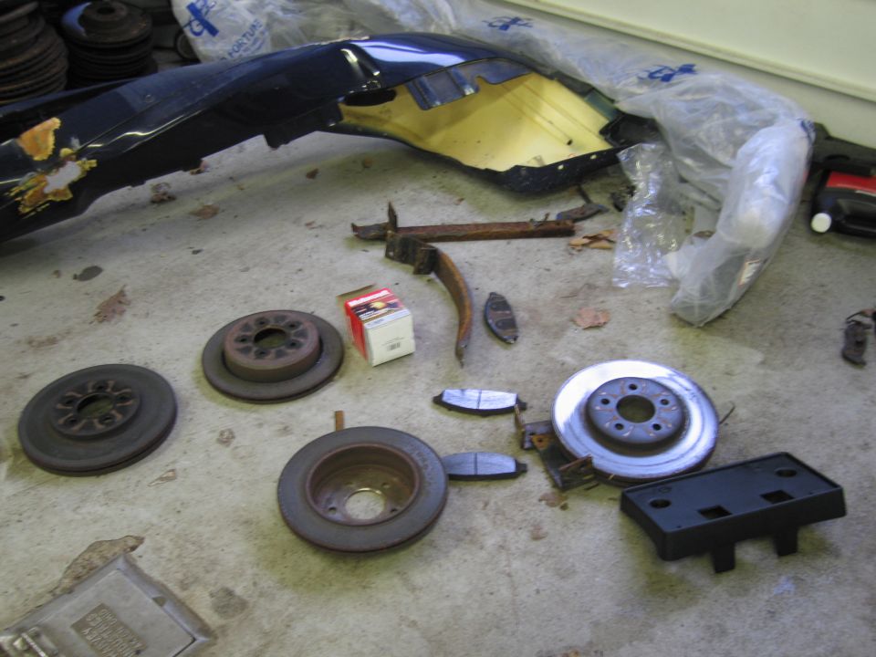 Car parts including brake rotors, brake pads, and a damaged blue bumper on a concrete floor