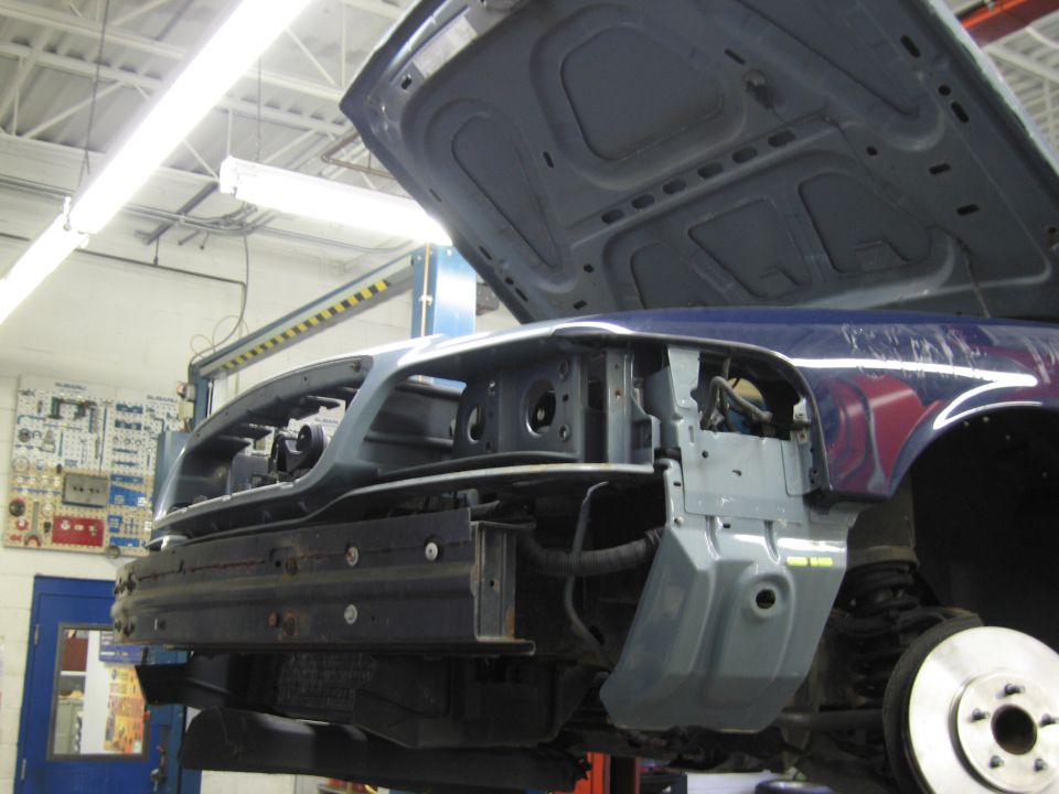 Car front end disassembled on a lift, hood open, showing internal components and bumper removed