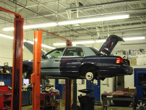 2001 Ford Crown Victoria - A two-tone blue and grey sedan is raised on a car lift in a garage, with its hood and trunk open for service.