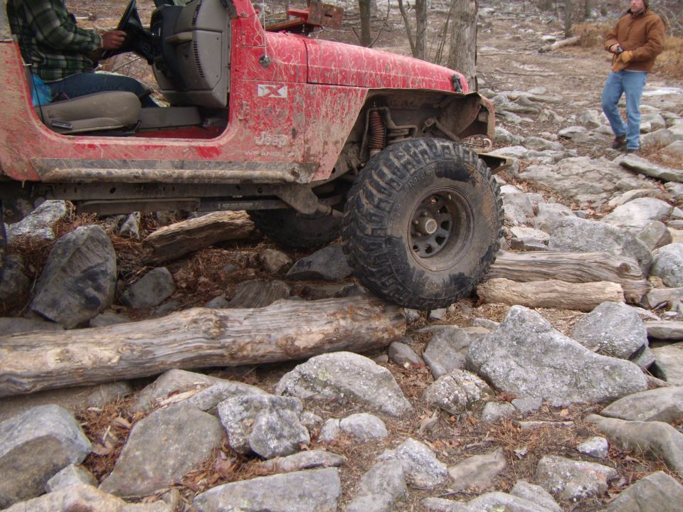 Red Jeep Wrangler on rocky terrain, driver visible, front right tire on log