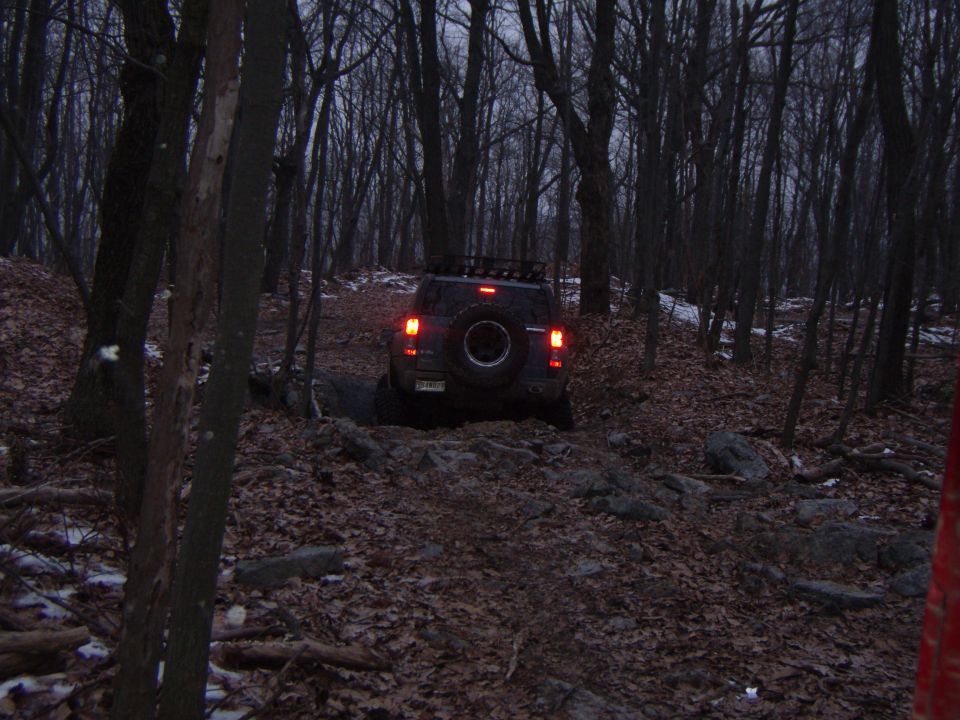 SUV with a roof rack and spare tire on the back drives on a rocky, leaf-covered trail through a dark, bare forest