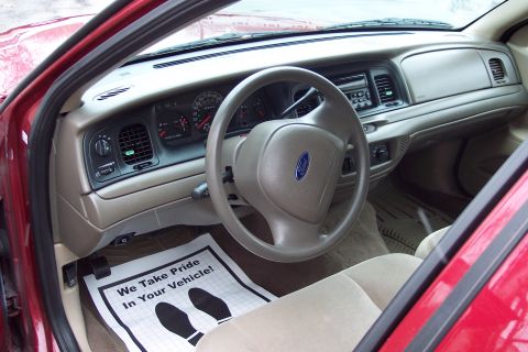 2001 Ford Crown Victoria - The interior of a red 2001 Ford Crown Victoria shows beige upholstery and a dashboard with a Ford logo on the steering wheel.