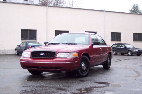 2001 Ford Crown Victoria - A red 2001 Ford Crown Victoria is parked in front of a building with other vehicles in the background.
