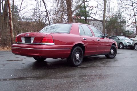 2001 Ford Crown Victoria - The rear view of a red sedan shows its taillights and chrome trim against a backdrop of trees and a cloudy sky.