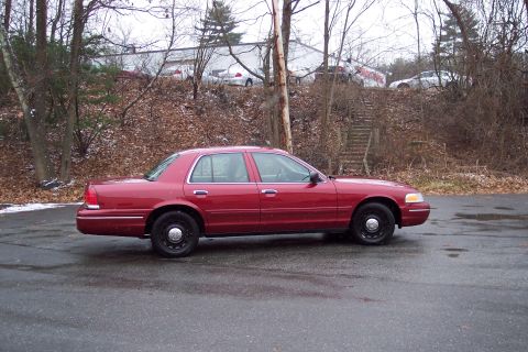 2001 Ford Crown Victoria - A red 2001 Ford Crown Victoria sedan is parked on a wet asphalt surface with trees and a building in the background.