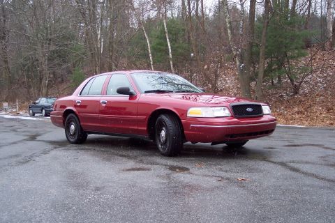 2001 Ford Crown Victoria - A red 2001 Ford Crown Victoria is parked on a wet road with trees in the background.